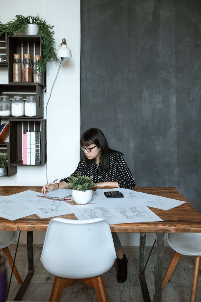 A woman intensely working at a wooden table in a modern office setting, surrounded by design plans and decor.