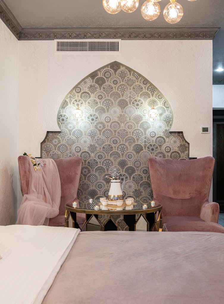 Stylish bedroom with comfortable bed and side table with set of cups and teapot placed between pink armchairs near ornamental wall